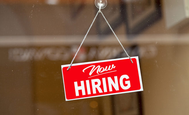 Red sign hanging at the glass door of a shop saying: "Now hiring".