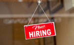 Red sign hanging at the glass door of a shop saying: "Now hiring".