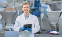 Young Male Quality Supervisor or Food Technician is Inspecting the Automated Production at a Dumpling Food Factory. Employee Uses a Tablet Computer for Work. He Looks to the Camera and Smiles.